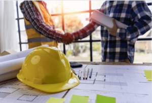 Screenshot-2023-04-06-123937 - Aryo Systems A yellow hard hat on a table with construction blueprints, as two people shake hands in the background, symbolizing a construction agreement or partnership.