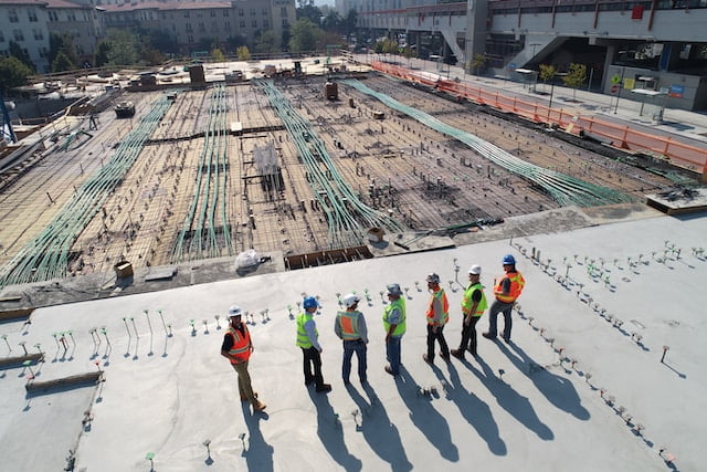 scott-blake-x-ghf9LjrVg-unsplash-1 - Aryo Systems A group of construction workers wearing safety gear standing on a large building site with visible rebar and materials.