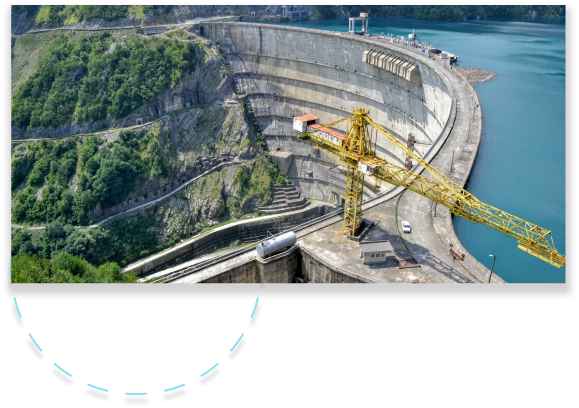 Construction assistance - Aryo Systems A massive concrete dam surrounded by lush green mountains and a reservoir, with construction equipment visible in the foreground.